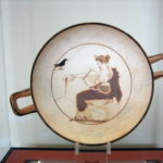 キリックスに描かれたアポロン　右手でワインを注ぎ、左手で竪琴を弾いている。服装、髪型が現代的にみえる。カラスは彼が愛したコロニスを表している。